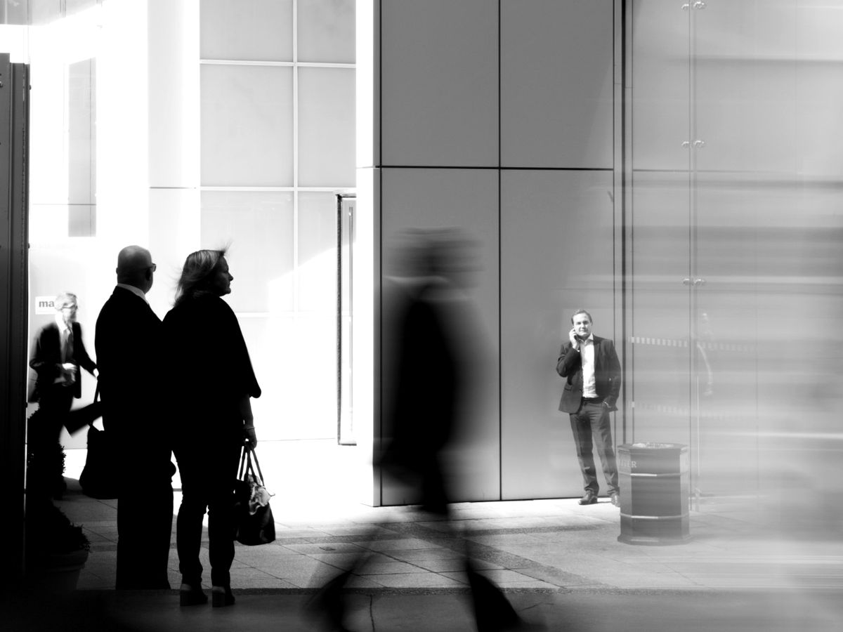 People on a city sidewalk outside a glass office building, one standing still on a phone call while others hurry past